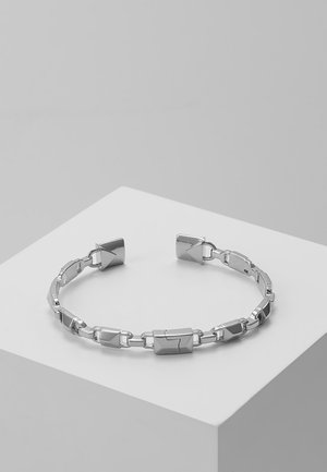 Bracelet à chaîne ouverte ton argent avec des maillons rectangulaires et carrés ornés de motifs de flèches gravées, présenté sur un socle blanc.