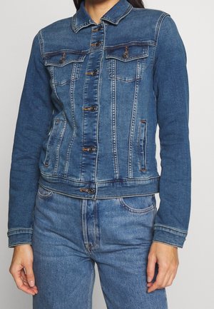 Kobieta ubrana w niebieską kurtkę dżinsową z zapięciami na guziki oraz pasujące niebieskie dżinsy na jednolitym tle.