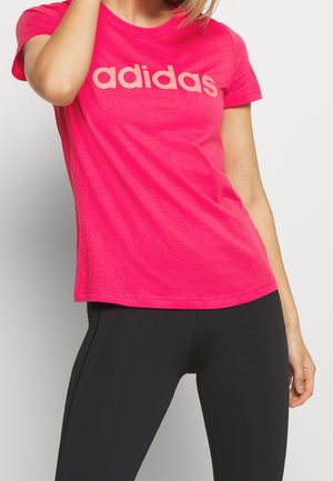 Femme portant un t-shirt Adidas rose vif et des leggings noirs, se tenant devant un fond clair uni, bras levé vers la tête.