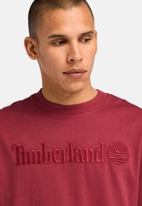 Camiseta de algodón marrón con cuello redondo, que presenta el logo bordado en relieve de "Timberland" y un emblema de árbol en el pecho. Textura suave.