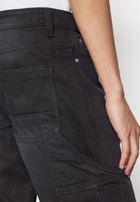 Pantalones de mezclilla negros con un acabado texturizado, que cuentan con múltiples bolsillos y un diseño ajustado, realzado por costuras visibles y herrajes metálicos.