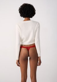 Perizoma rosso in pizzo con bordi arricciati e un dettaglio ritagliato circolare, indossato con un maglione bianco aderente a maniche lunghe. Carnagione liscia.