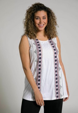 Top bianco senza maniche con ricami intricati in rosso e blu lungo la parte anteriore. Tessuto morbido, vestibilità ampia e orlo curvo. Abbinato a pantaloni neri.