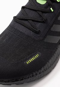 Zbliżenie czarnego obuwia biegowego Adidas z czarnymi sznurowadłami, teksturowaną cholewką i żółtą etykietą "AEROREADY" po boku.