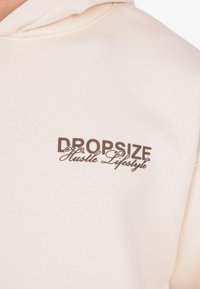 Zbliżenie na kremowy hoodie z brązowym napisem "DROPSIZE Hustle Lifestyle" na klatce piersiowej.
