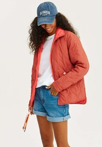 Kurtka pikowana w kolorze koralowym z kontrastowym pomarańczowym zamkiem, noszona na białym podkoszulku i szortach dżinsowych. Niebieska czapka z napisem "LOS ANGELES".