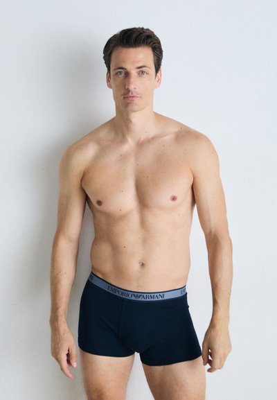 Sorte boxer briefs med grå "Emporio Armani" elastikkant. Stoffet ser glatt ut, med en tettsittende design. Modellen har et nøytralt uttrykk.