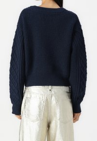 Granatowy sweter z prążkowanym wzorem i teksturowanymi rękawami w splot kablowy, z okrągłym dekoltem, w połączeniu z błyszczącymi srebrnymi spodniami. Neutralne tło.