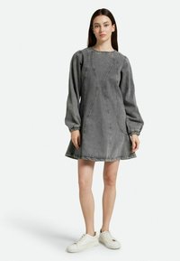 Vestido de mezclilla gris con mangas largas abullonadas, detalles en las costuras y un dobladillo acampanado. Lleva puestas zapatillas blancas, contra un fondo liso.