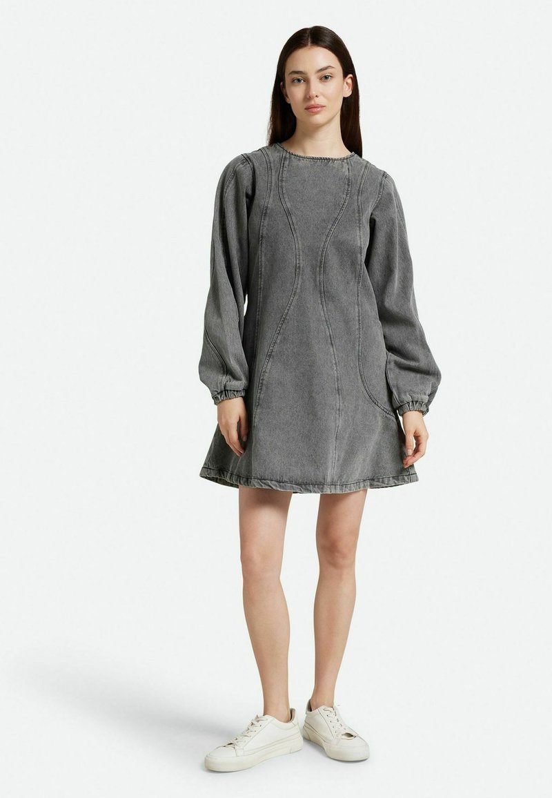 Vestido de mezclilla gris con mangas largas abullonadas, detalles en las costuras y un dobladillo acampanado. Lleva puestas zapatillas blancas, contra un fondo liso.