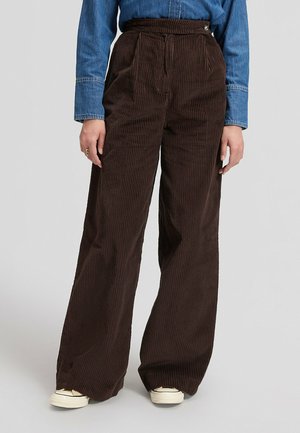 Persona che indossa pantaloni marroni a gamba larga in velluto a coste, camicia di jeans blu e sneakers bianche, in piedi contro uno sfondo chiaro e semplice.