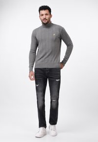 Grauer gerippter Rollkragenpullover mit gelbem Logo, kombiniert mit distressed schwarzen Jeans und weißen Sneakers. Schlichte, moderne Silhouette.