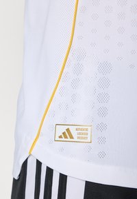 Hvid atletisk skjorte med gule accenter, mesh tekstur, buet kant og guld Adidas mærke med "AUTENTISK LICENSERET PRODUKT."