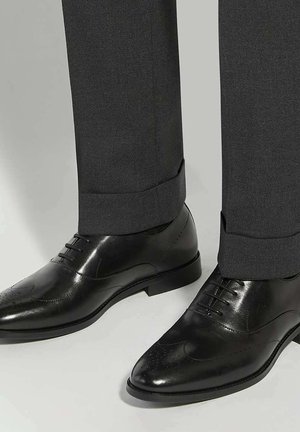Chaussures de ville noires vernies avec détails à bout golf, portées avec un pantalon formel gris foncé.