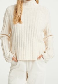 Pull crème côtelé à col montant et manches longues, présentant un motif tricoté texturé et une coupe décontractée, associé à un pantalon blanc côtelé.