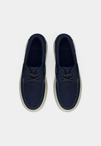 Παπούτσια από σουέτ σε ναυτικό μπλε χρώμα με χαμηλό προφίλ, στρογγυλή μύτη, αντίθετη λευκή ραφή και καφέ κορδόνια. Απαλή υφή σε όλη την επιφάνεια.