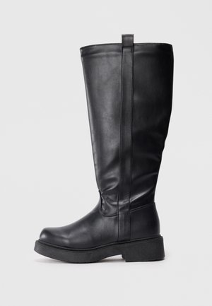 Botte en cuir noire montant jusqu'au genou avec une semelle épaisse et un onglet de tirage en haut, montrée de côté sur un fond neutre.