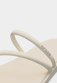 Μπεζ σάνδαλο Crocs με δύο λεπτές τιράντες, η μία φέρει το λογότυπο «crocs» με μικρά, ανάγλυφα γράμματα.