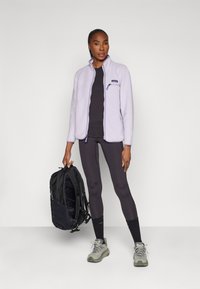 Fleece jack met een lichte lavendelkleur, hoge kraag en een zak met rits, gecombineerd met zwarte legging, grijze wandelschoenen en een zwarte rugzak.