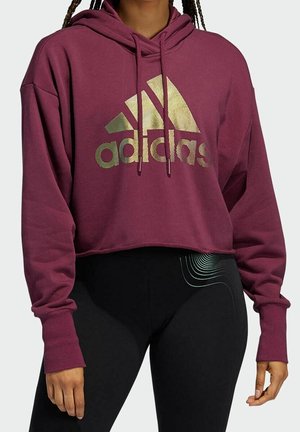 Femme portant un sweat à capuche court Adidas bordeaux avec un logo doré et des leggings noirs ornés d'un motif de ligne blanche incurvée sur la hanche.