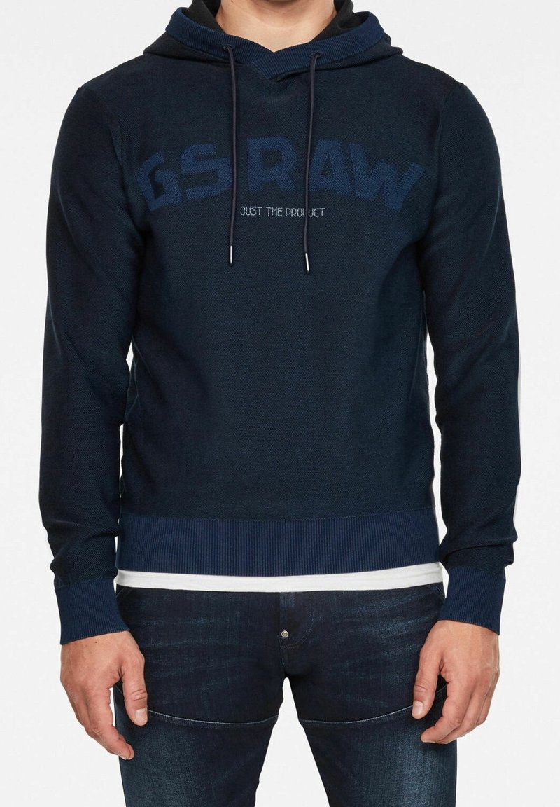 Homme portant un sweat à capuche bleu marine avec les inscriptions "G S RAW" et "JUST THE PRODUCT", associé à un jean bleu foncé et un débardeur blanc.