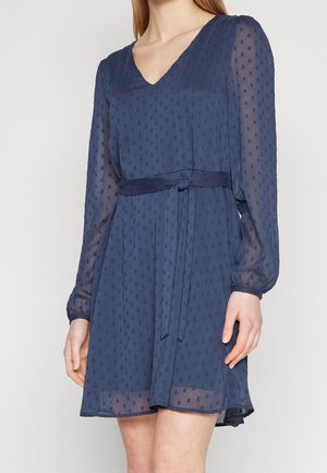 Femme portant une robe bleu marine longueur genoux avec des points texturés, des manches longues transparentes, un décolleté en V et une ceinture nouée à la taille.