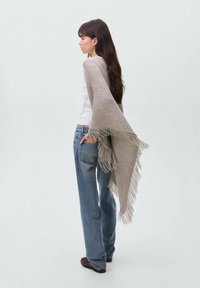 Gray knit shawl with fringe edges worn over a white top and blue jeans, displayed from the side against a light background.