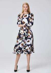 Femme portant une robe à manches longues et mi-longue avec des motifs abstraits multicolores marron, blanc, noir et lavande, se tenant les bras croisés.