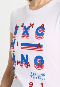 Bílý bavlněný tričko s výrazným, multikulturním textem v červené a modré barvě, který má různorodé textury a vzory. Mezi pozoruhodné designové prvky patří vrstvená písmena a pruhy.