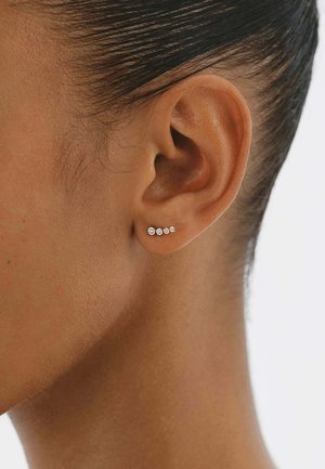 Primer plano de una oreja de piel morena con un pequeño pendiente plateado que presenta cinco diamantes redondos ascendentes a lo largo del lóbulo.