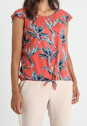 Femme portant un haut corail à manches courtes avec des motifs de feuilles bleues et vertes, noué à la taille, assorti à un pantalon beige clair.
