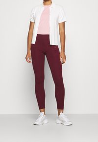 Leggings burdeos con cintura elástica alta combinados con una camiseta rosa claro y una camisa blanca de botones. Zapatillas deportivas blancas con suelas texturizadas.