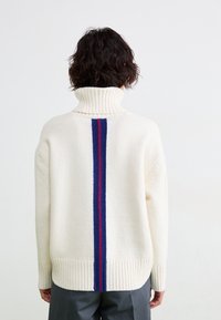 Cremefarvet sweater med høj krave. Har en vertikal blå og rød stribe på ryggen. Strikket med tekstur og ribkant ved sømmen og ærmerne.