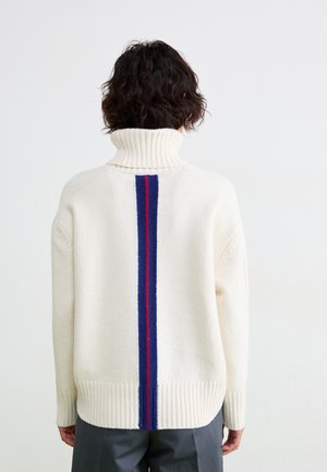 Kremfarget genser med høy krage. Har en vertikal blå og rød stripe på baksiden. Strukturert strikk med ribbestrikket kant og mansjetter.