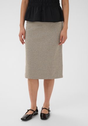 Femme portant une jupe beige à carreaux longueur genou, un haut noir et des chaussures Mary Jane noires vernies, debout devant un fond uni.