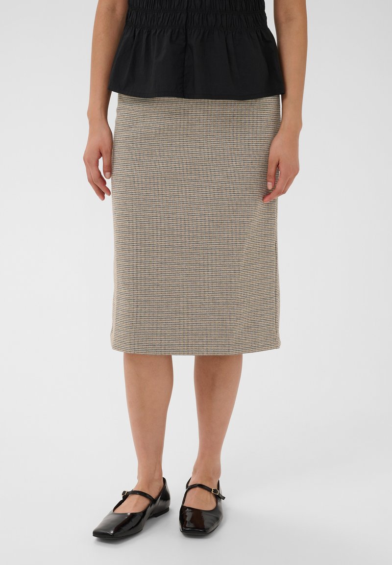 Femme portant une jupe beige à carreaux longueur genou, un haut noir et des chaussures Mary Jane noires vernies, debout devant un fond uni.