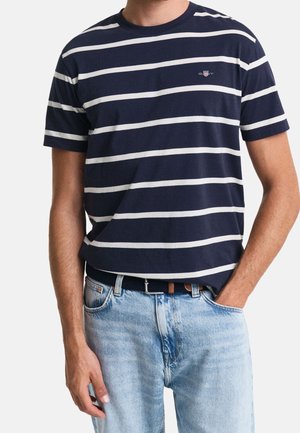 Homme portant un t-shirt à manches courtes à rayures horizontales bleu marine et blanc, rentré dans un jean bleu clair, avec une main dans la poche.
