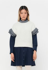 Frau, die einen weißen kurzärmeligen Strickpullover über einem navyblauen langärmeligen plissierten Kleid mit gestreiften Rüschendetails trägt.