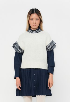 Woman wearing a white short-sleeve knit sweater over a navy blue long-sleeve pleated dress with striped ruffle details.