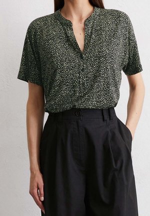 Vrouw draagt een zwart shirt met korte mouwen en witte spikkels, ingestopt in een zwarte broek met hoge taille, plooien en een knoopsluiting.