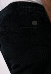 Pantalons en velours côtelé noir avec une texture côtelée, une taille élastique et un petit patch logo sur la poche arrière.