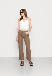 Top cropped branco canelado com um recorte em forma de coração, calças largas castanhas e sandálias pretas com tiras, em fundo claro e neutro.