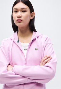 Νέα γυναίκα με ίσια σκούρα μαλλιά, φοράει ανοιχτό ροζ φούτερ με φερμουάρ πάνω από λευκό τοπ, με σταυρωμένα χέρια και ουδέτερη έκφραση, σε απλό φόντο.