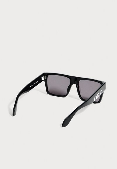 Lunettes de soleil rectangulaires noires avec des verres foncés et un texte blanc sur la branche gauche, présentées sur un fond gris clair.