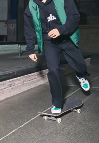 Skater noszący zieloną puchową kamizelkę na czarnej bluzie z kapturem, ciemne spodnie i trampki z zielonymi akcentami, jeżdżący na deskorolce w skateparku.