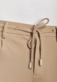 Beżowe spodnie z plecionym sznurkiem ściągającym zawiązanym w kokardę na pasku, noszone z włożoną białą koszulą.