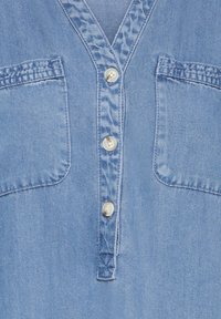 Haut en denim bleu clair avec un col en V, une fermeture à quatre boutons et deux poches poitrine avec détails de couture. Texture lisse sur toute la surface.