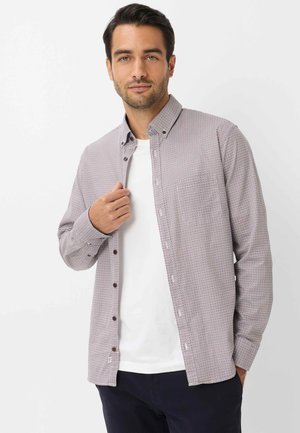 Mann mit dunklen Haaren, der ein gemustertes Hemd über einem weißen T-Shirt und dunkler Hose trägt, steht vor einem schlichten weißen Hintergrund.