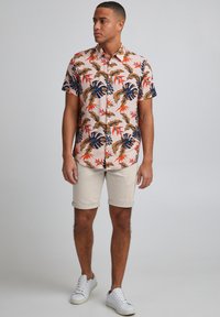 Camicia a maniche corte color rosa chiaro con motivo di foglie tropicali in verde, blu navy e rosso, abbinata a pantaloncini beige risvoltati e scarpe da ginnastica bianche.