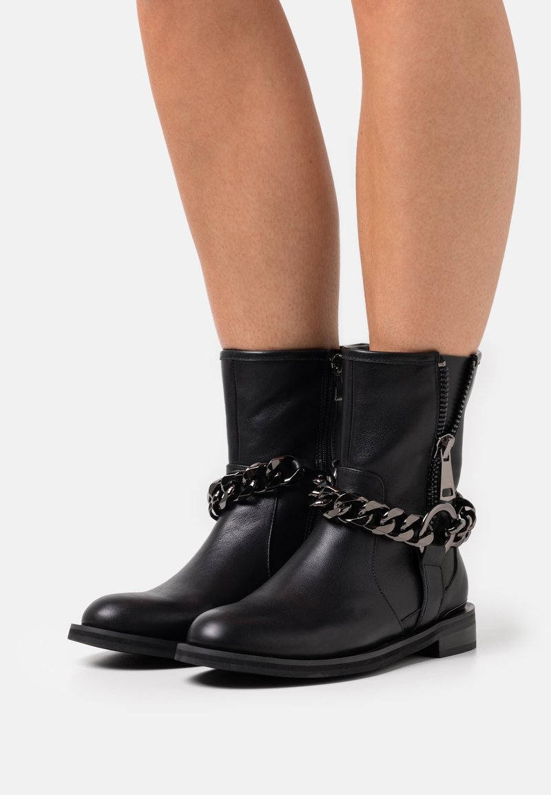 Botines negros de cuero con un acabado texturizado, que cuentan con una cremallera lateral y un acento de cadena metálica gruesa. Punta cuadrada y tacón bajo.
